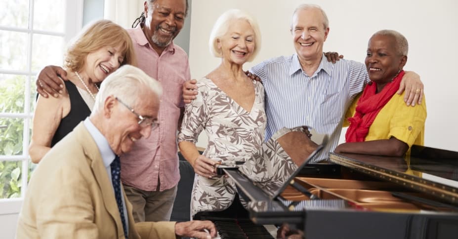 Happy seniors playing piano with friends gathered around them.